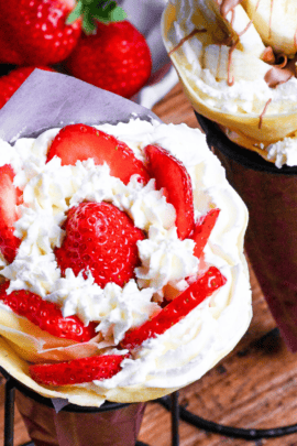 Two Japanese-style crepes filled with cream and strawberries, and cream and bananas wrapped with maroon paper and held in a black wire stand on a wooden background