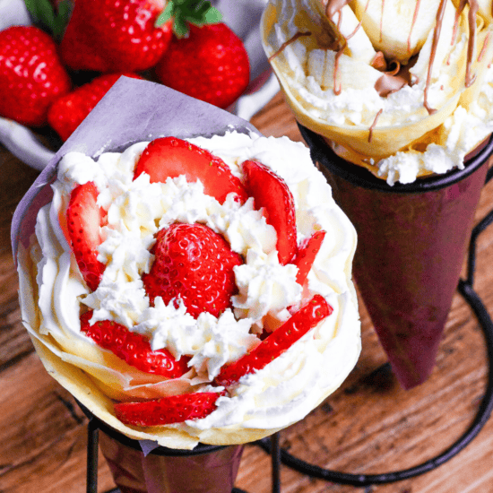 Two Japanese-style crepes filled with cream and strawberries, and cream and bananas wrapped with maroon paper and held in a black wire stand on a wooden background