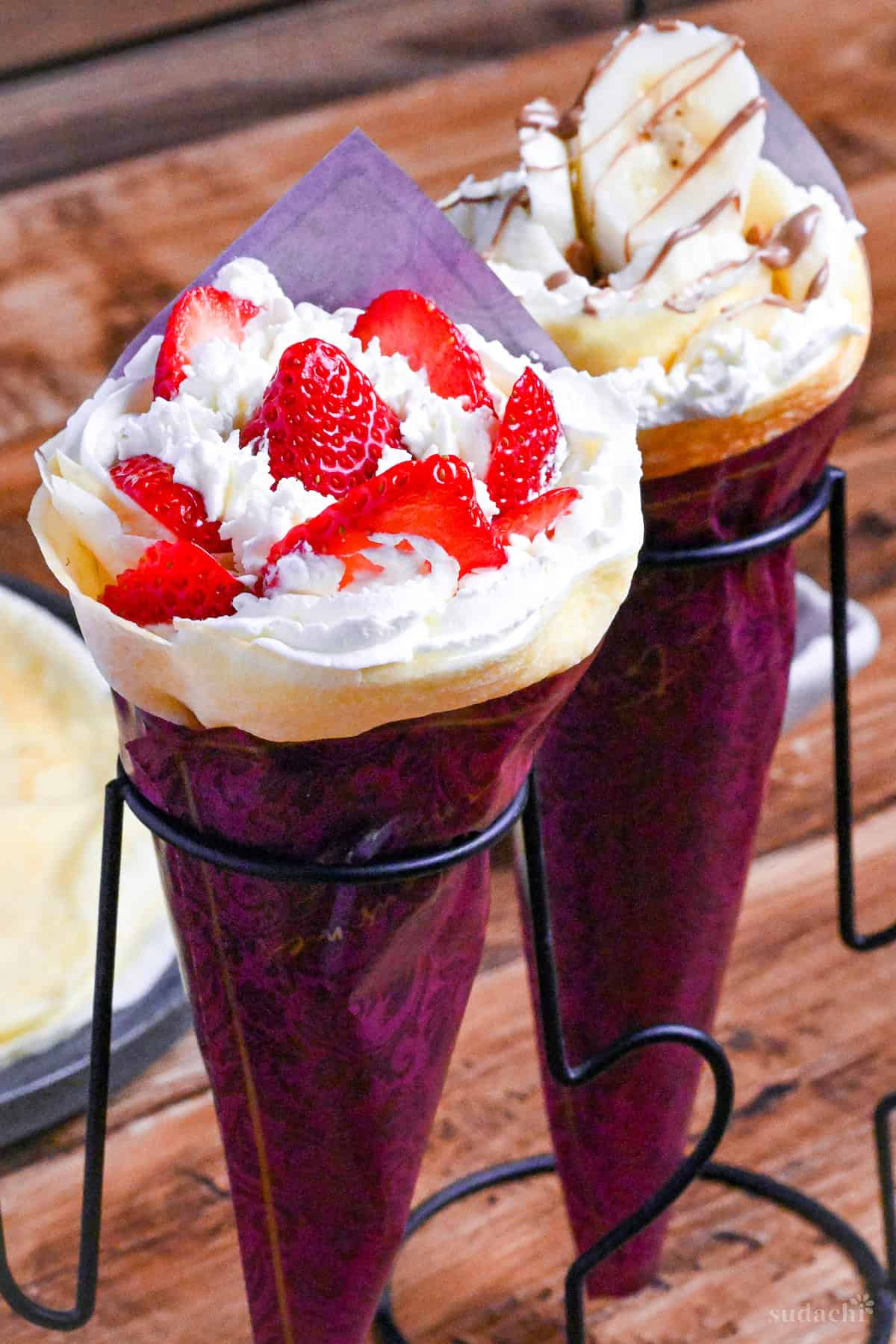 Two Japanese-style crepes filled with cream and strawberries, and cream and bananas wrapped with maroon paper and held in a black wire stand on a wooden background