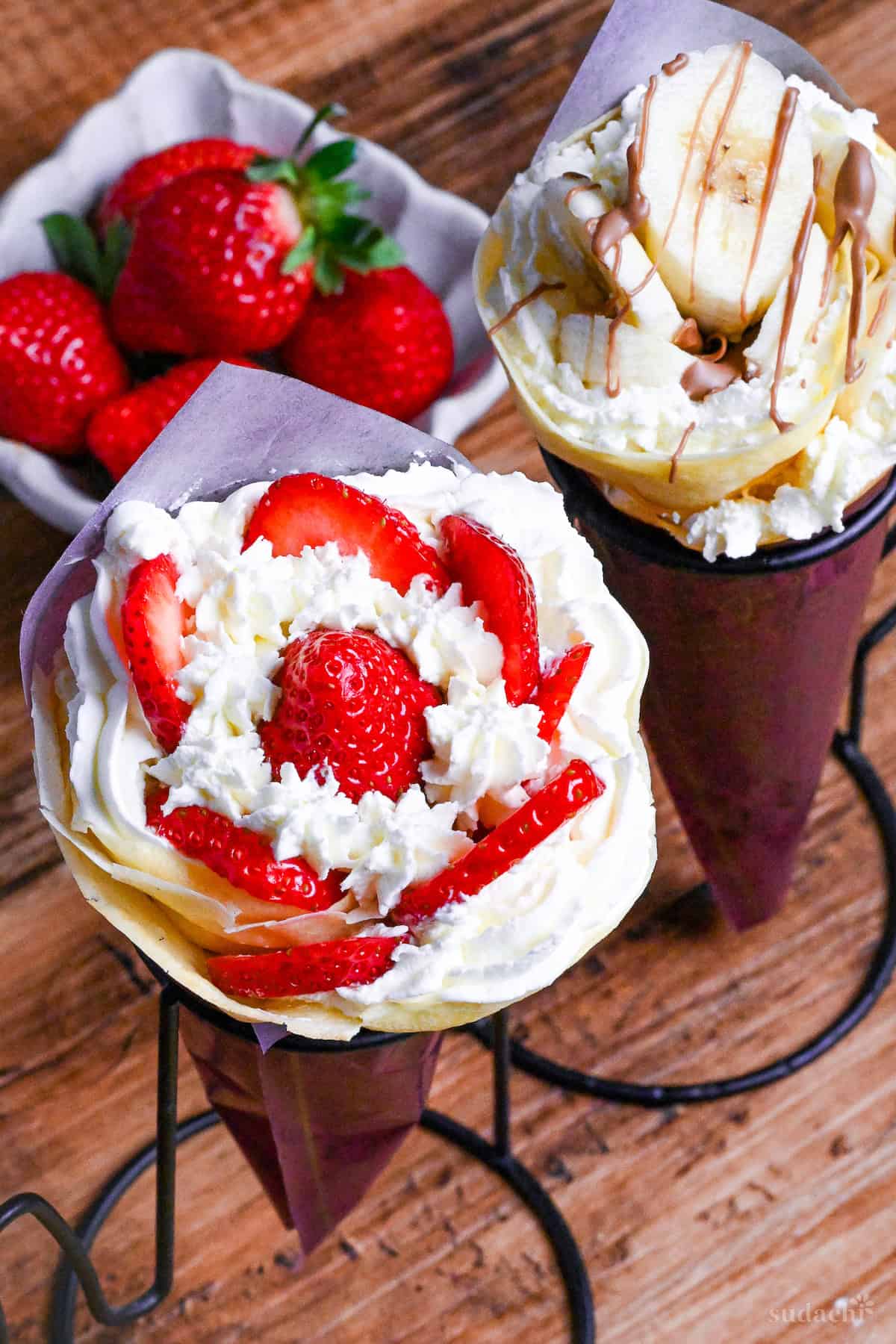 Two Japanese-style crepes filled with cream and strawberries, and cream and bananas wrapped with maroon paper and held in a black wire stand on a wooden background