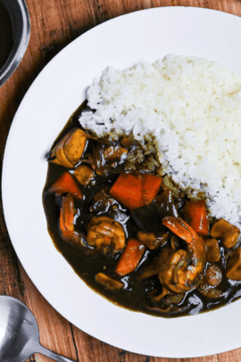 Black squid ink seafood curry and rice on a white plate next to a steel gravy boat and black soup spoon on a wooden table top down view