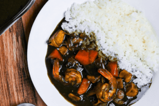 Black squid ink seafood curry and rice on a white plate next to a steel gravy boat and black soup spoon on a wooden table top down view