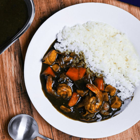 Black squid ink seafood curry and rice on a white plate next to a steel gravy boat and black soup spoon on a wooden table top down view