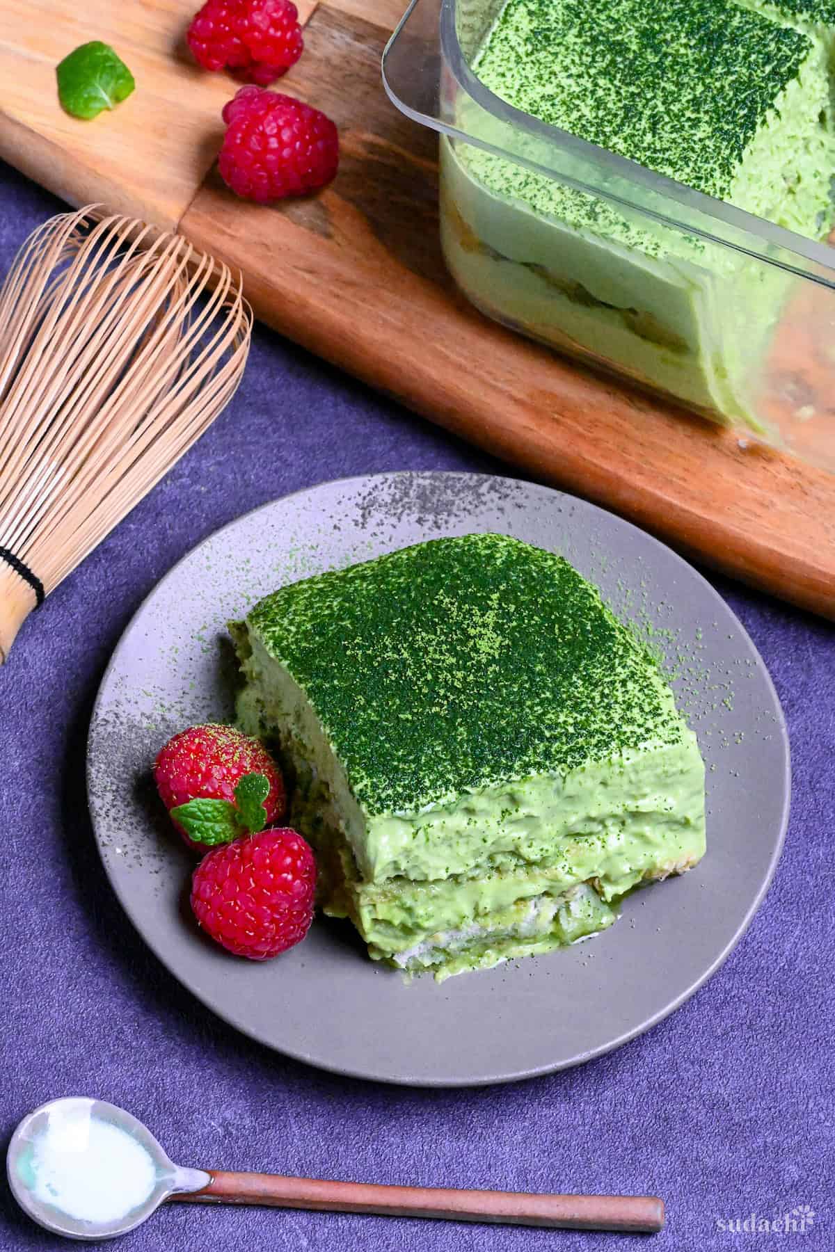 A square piece of matcha tiramisu on a gray plate with two raspberries and a mint leaf with matcha whisk in the background