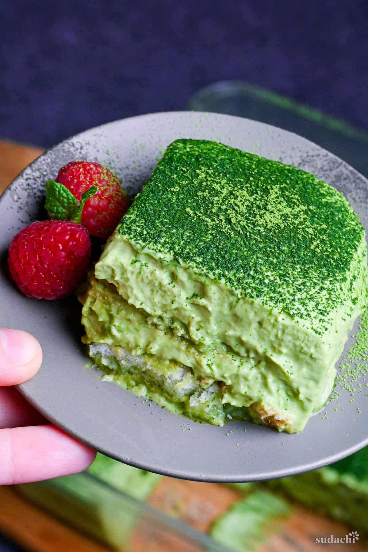 A square piece of matcha tiramisu on a gray plate with two raspberries and a mint leaf