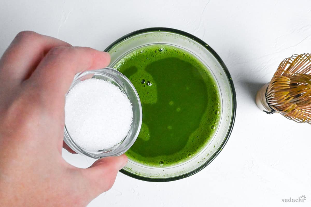 pouring sugar into matcha and water mixture in a glass bowl