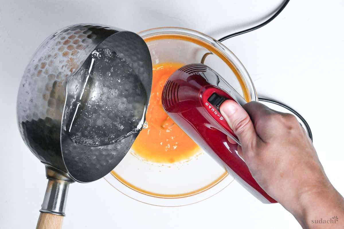 pouring sugar syrup into egg yolks in a glass mixing bowl