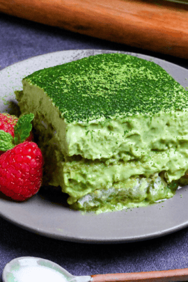 A square piece of matcha tiramisu on a gray plate with two raspberries and a mint leaf