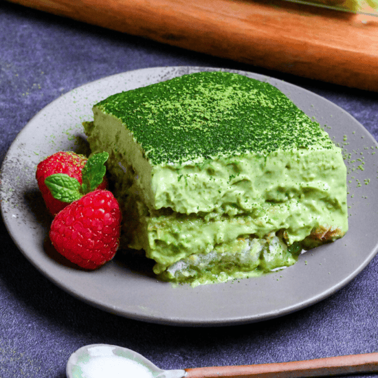 A square piece of matcha tiramisu on a gray plate with two raspberries and a mint leaf