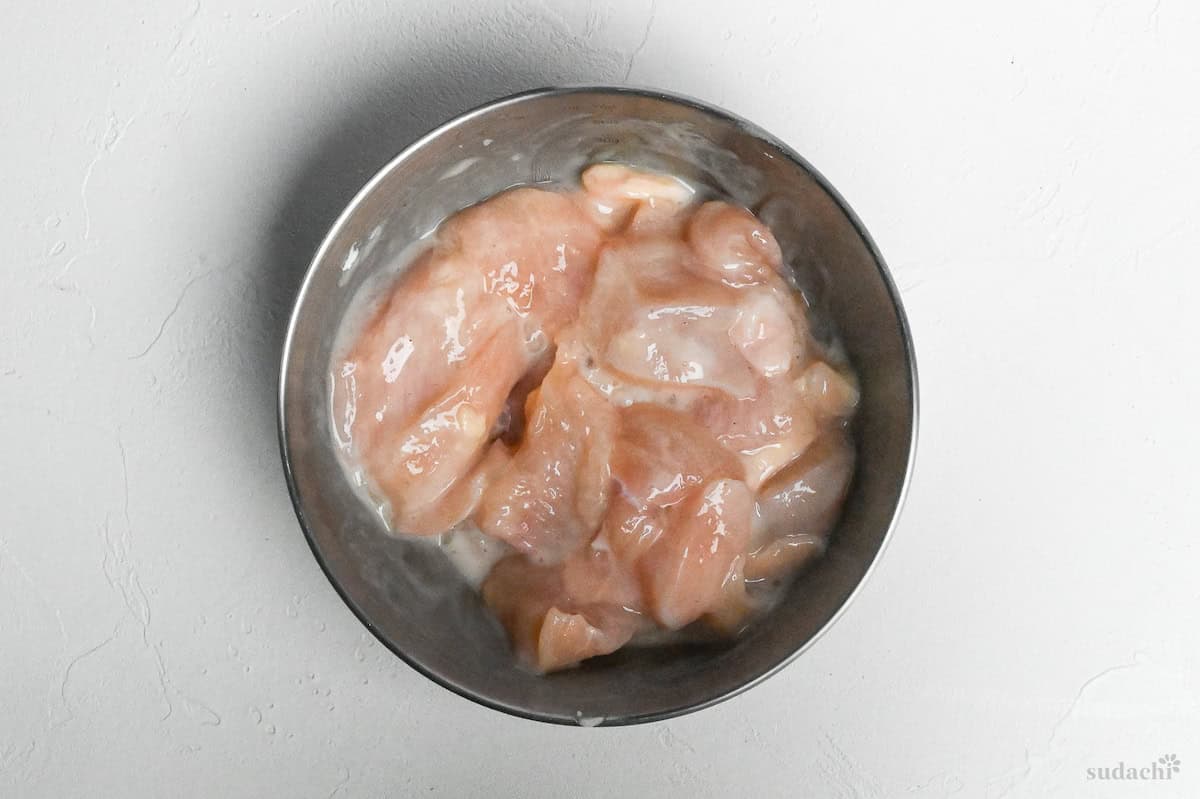 Pieces of chicken breast in a steel mixing bowl coated with egg whites and potato starch to tenderize