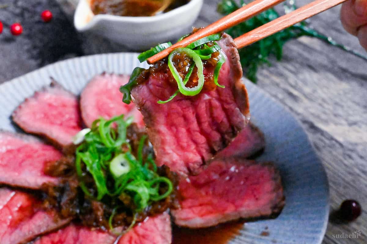 A slice of Japanese style roast beef cooked rare held up with wooden chopsticks