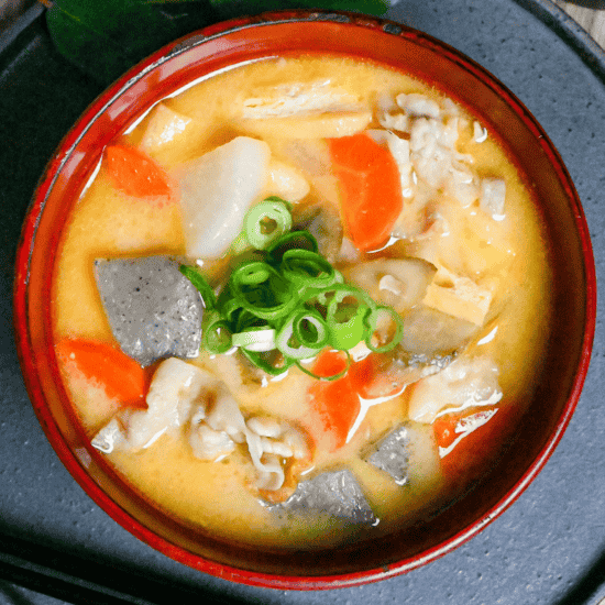 Sake lees soup (kasujiru) made with pork, vegetables and konnyaku in a red bowl on a gray ceramic placemat