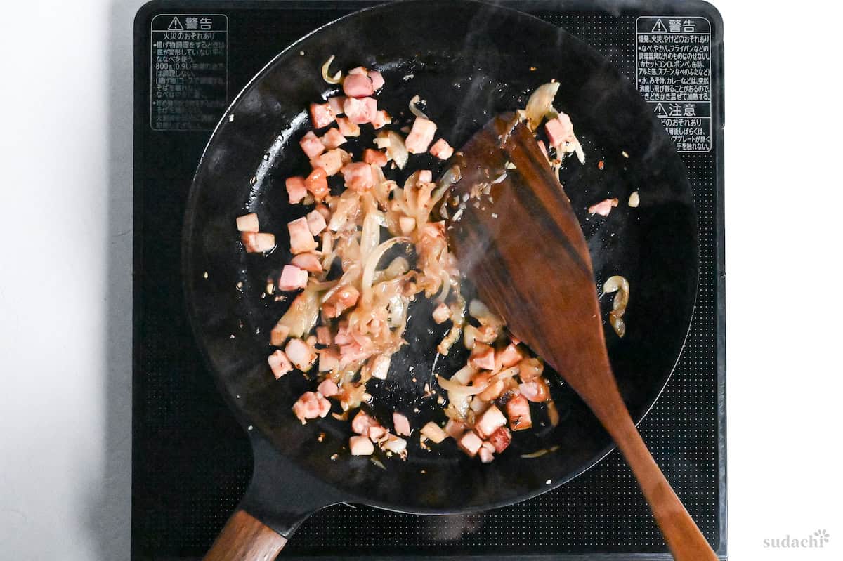 bacon and sliced onions frying in a pan on the stove with a wooden spatula