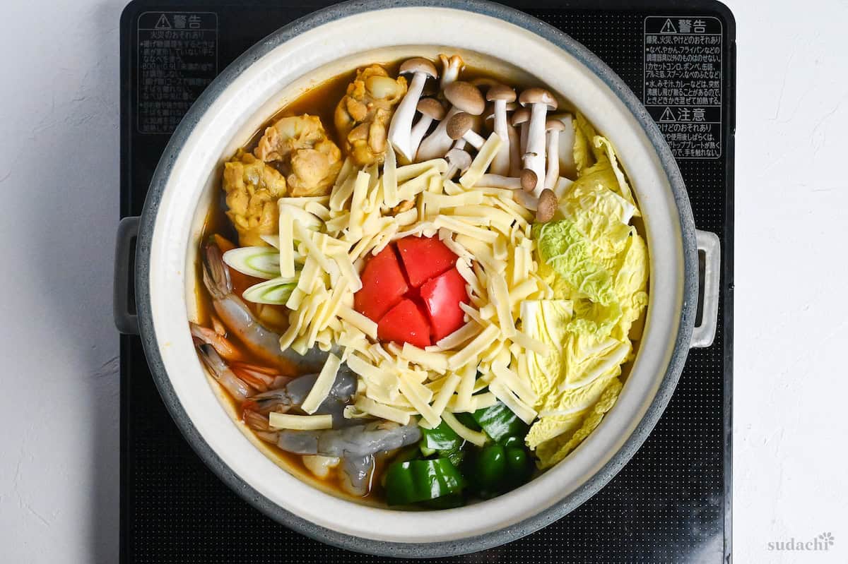 Japanese curry hot pot topped with a tomato, cheese, chicken, prawns and various vegetables on the a black stove on a white background