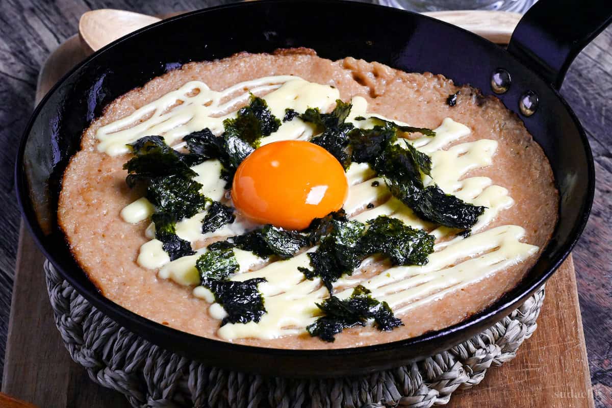 side view of fluffy grilled grated Japanese yam topped with mayonnaise, nori and an egg yolk in a black pan on a wooden chopping board