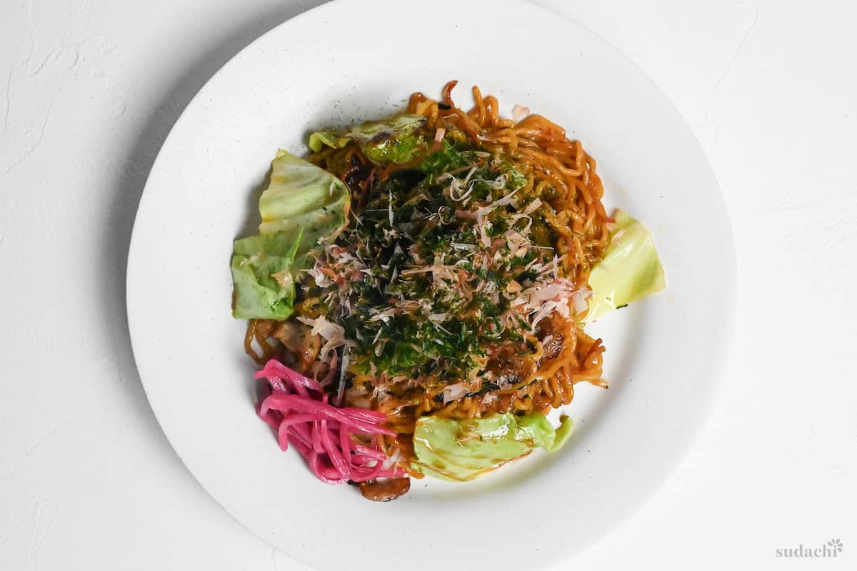 Authentic Japanese yakisoba on a white plate garnished with aonori, bonito flakes and pink pickled ginger (benishoga) on a white background