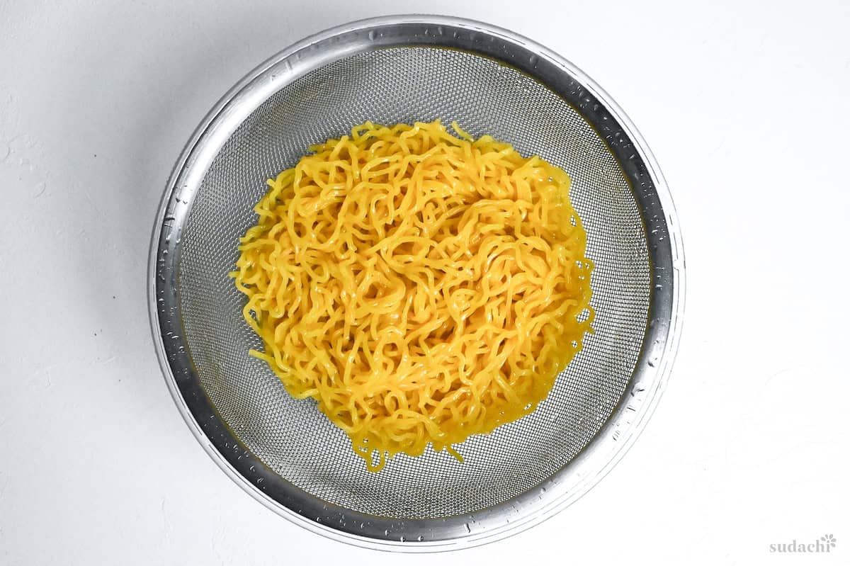 drained ramen noodles in a sieve over a glass bowl on a white background