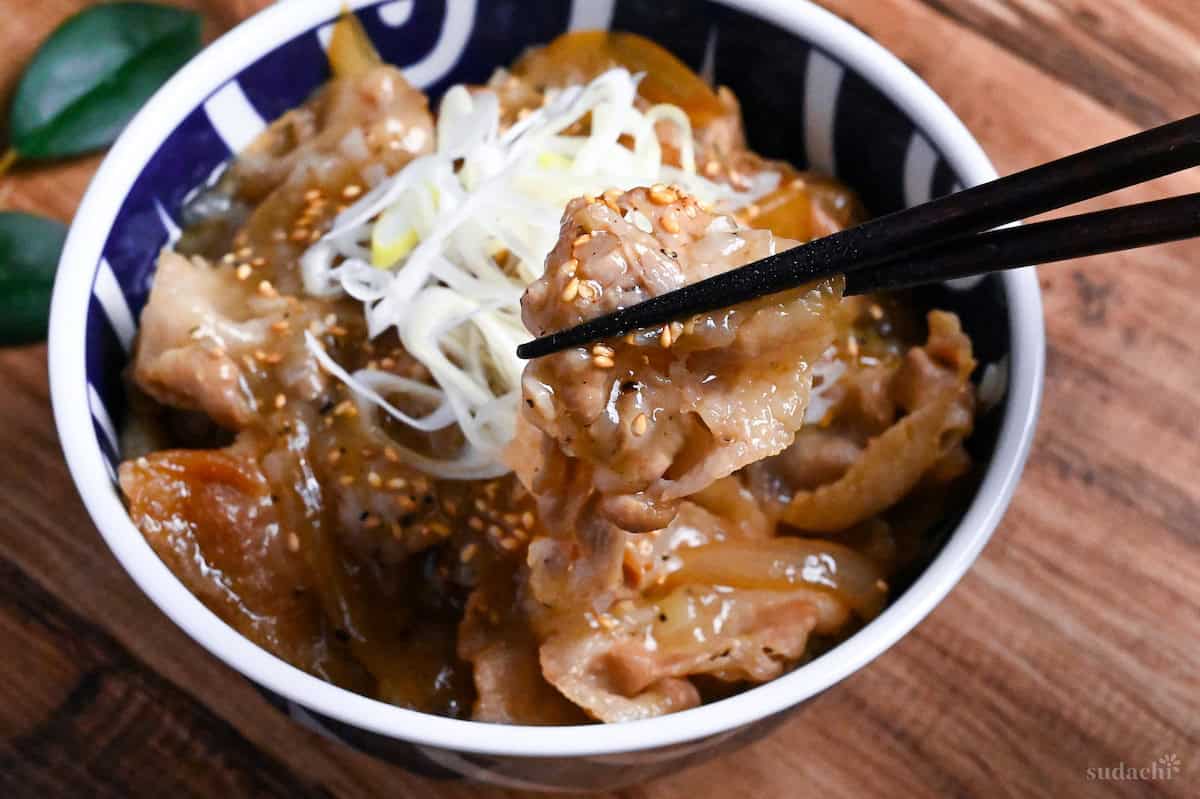 Yoshinoya Copycat Negi Shio Butadon topped with chopped Japanese leeks and sesame seeds in a white and blue bowl on a wooden surface with black chopsticks holding up a piece of thinly sliced pork