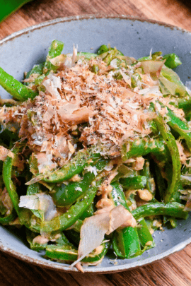 Mugen Piman (Addictive Bell Pepper) garnished with bonito flakes in a gray/blue bowl on wooden surface with green bell pepper in the background close up thumbnail