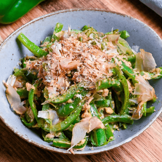 Mugen Piman (Addictive Bell Pepper) garnished with bonito flakes in a gray/blue bowl on wooden surface with green bell pepper in the background close up thumbnail