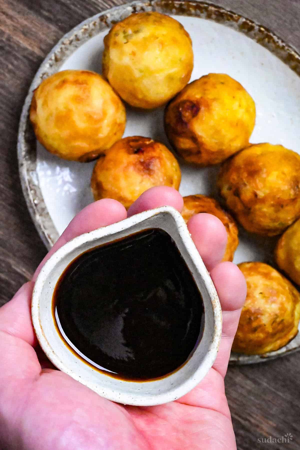 homemade takoyaki sauce in a small cream ceramic jug being held over bare takoyaki