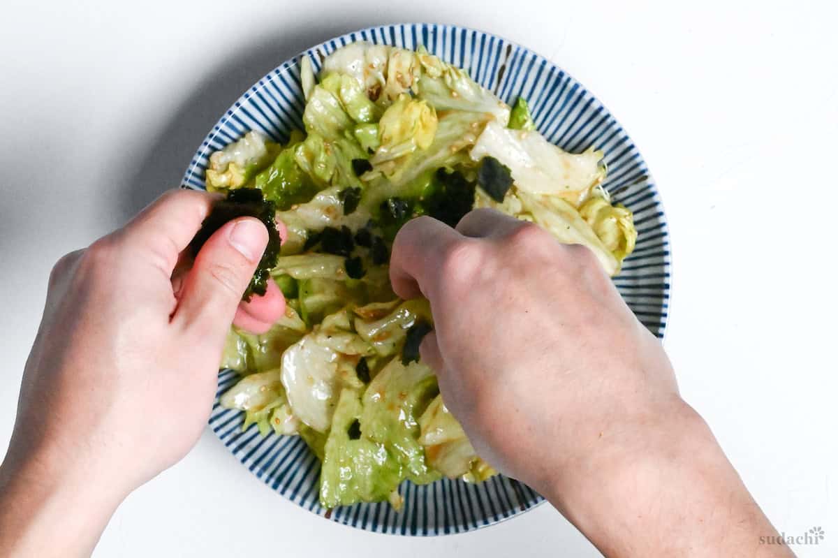 sprinkling torn nori over yamitsuki lettuce on a white and blue striped bowl