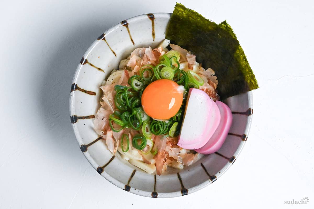 abura udon topped with bonito flakes, chopped green onions, nori, kamaboko fishcakes and a raw egg yolk served in an off-white bowl with brown stripes on a white background