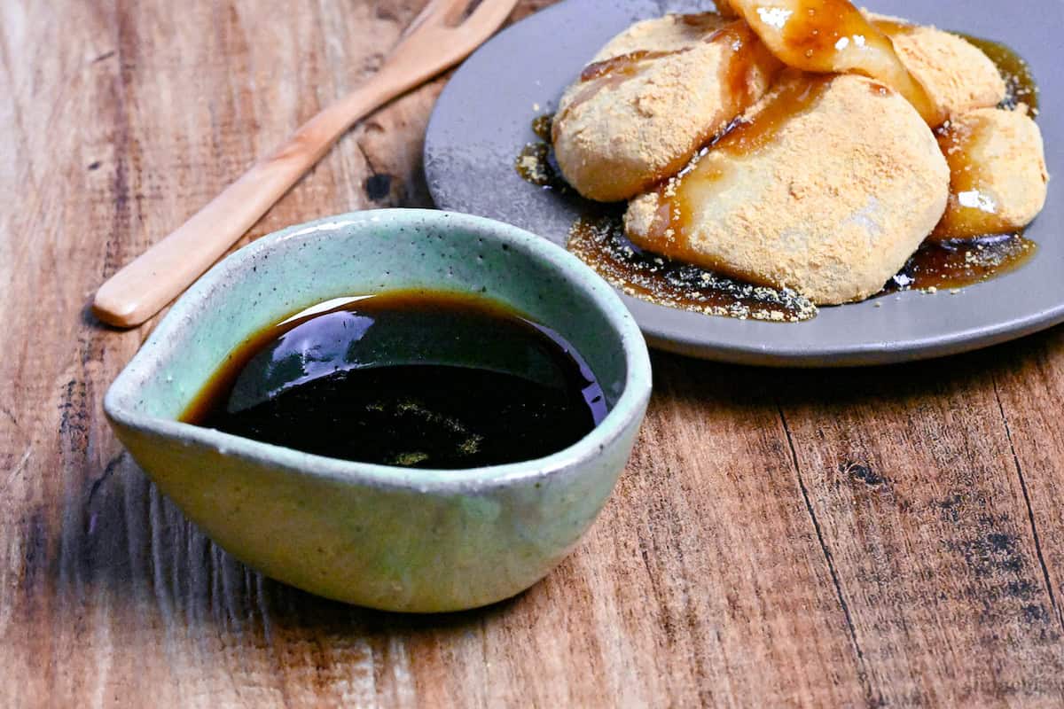 Homemade kuromitsu (Japanese dark sugar syrup) in a small green ceramic jug with it drizzled over mochi in the background