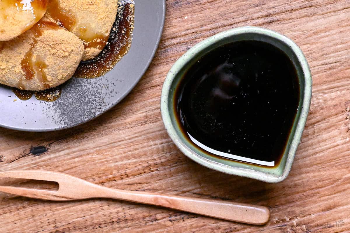 Homemade kuromitsu (Japanese dark sugar syrup) in a small green ceramic jug with it drizzled over mochi in the background top down