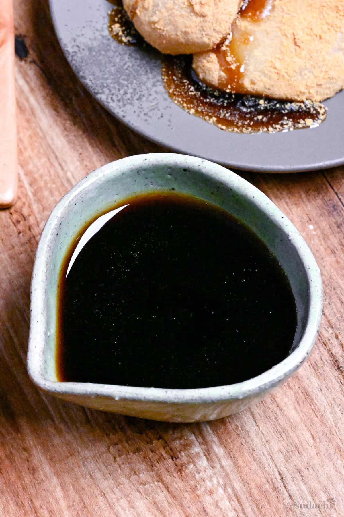 Homemade kuromitsu (Japanese dark sugar syrup) in a small green ceramic jug with it drizzled over mochi in the background