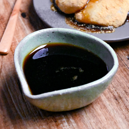 Homemade kuromitsu (Japanese dark sugar syrup) in a small green ceramic jug with it drizzled over mochi in the background