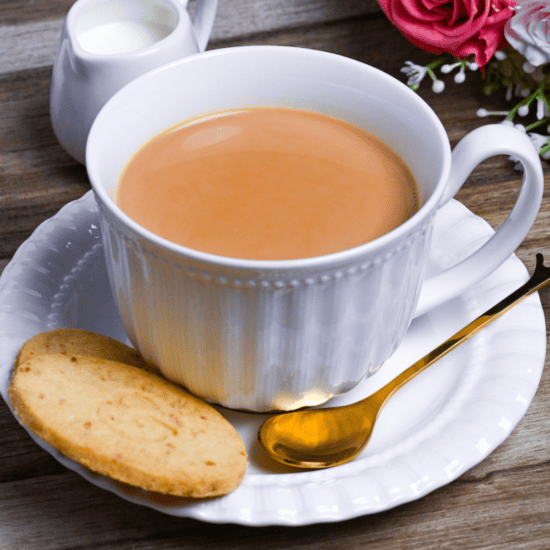 Japanese royal milk tea in a white cup with saucer, shortbread cookies and a golden teaspoon on a light wooden background with sugar cubes, a milk jug and flowers in the background