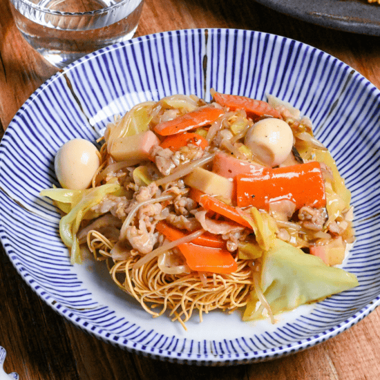 Sara udon served on a white and blue striped plate on a wooden surface next to black chopsticks and a glass of water