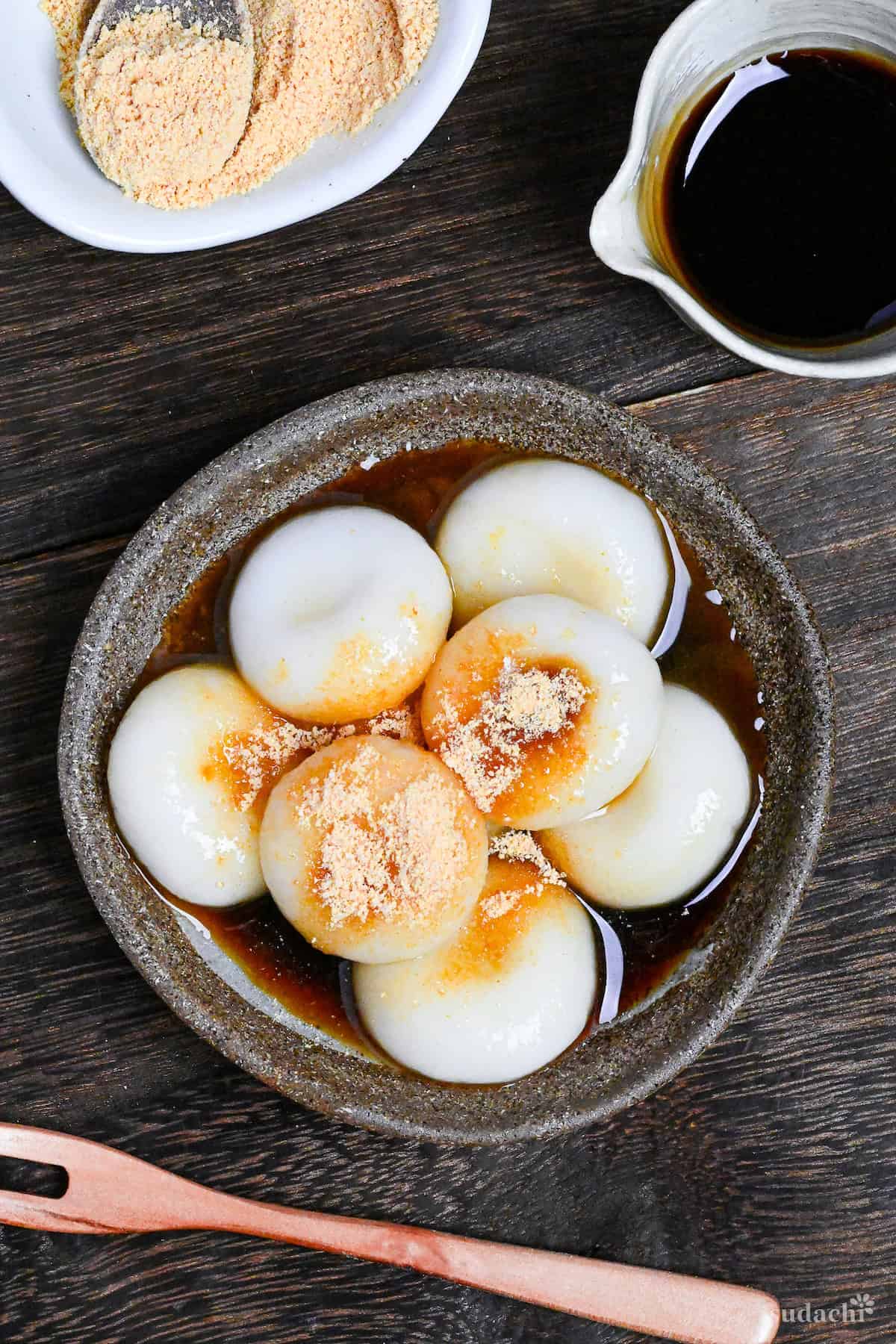 Shiratama dango (mochi balls) topped with kuromitsu (brown sugar syrup) and kinako (soybean powder) in a brown dish on a wooden table