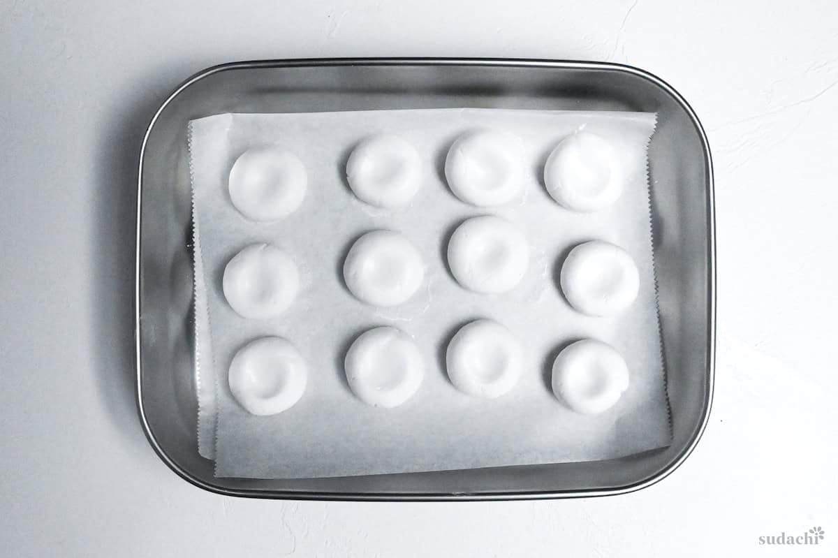 12 shaped (uncooked) shiratama dango on a baking sheet in a steel container on a white background