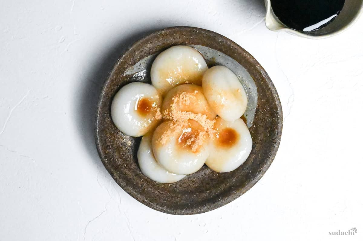 Shiratama dango (mochi balls) topped with kuromitsu (brown sugar syrup) and kinako (soybean powder) in a brown dish on a white background