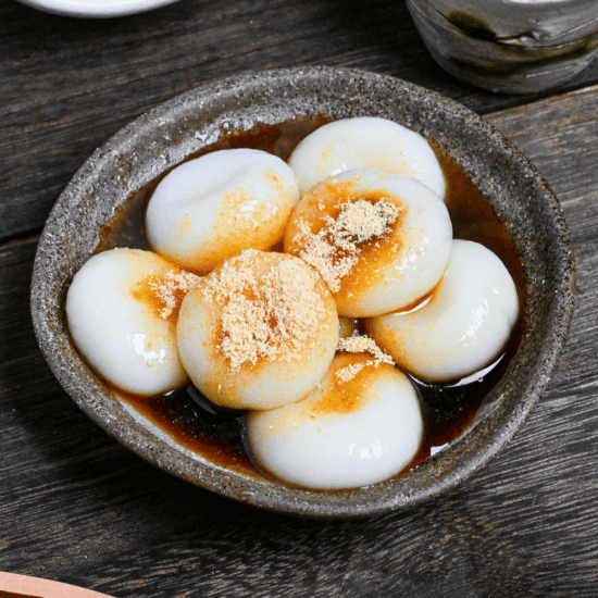 Shiratama dango (mochi balls) topped with kuromitsu (brown sugar syrup) and kinako (soybean powder) in a brown dish on a wooden table