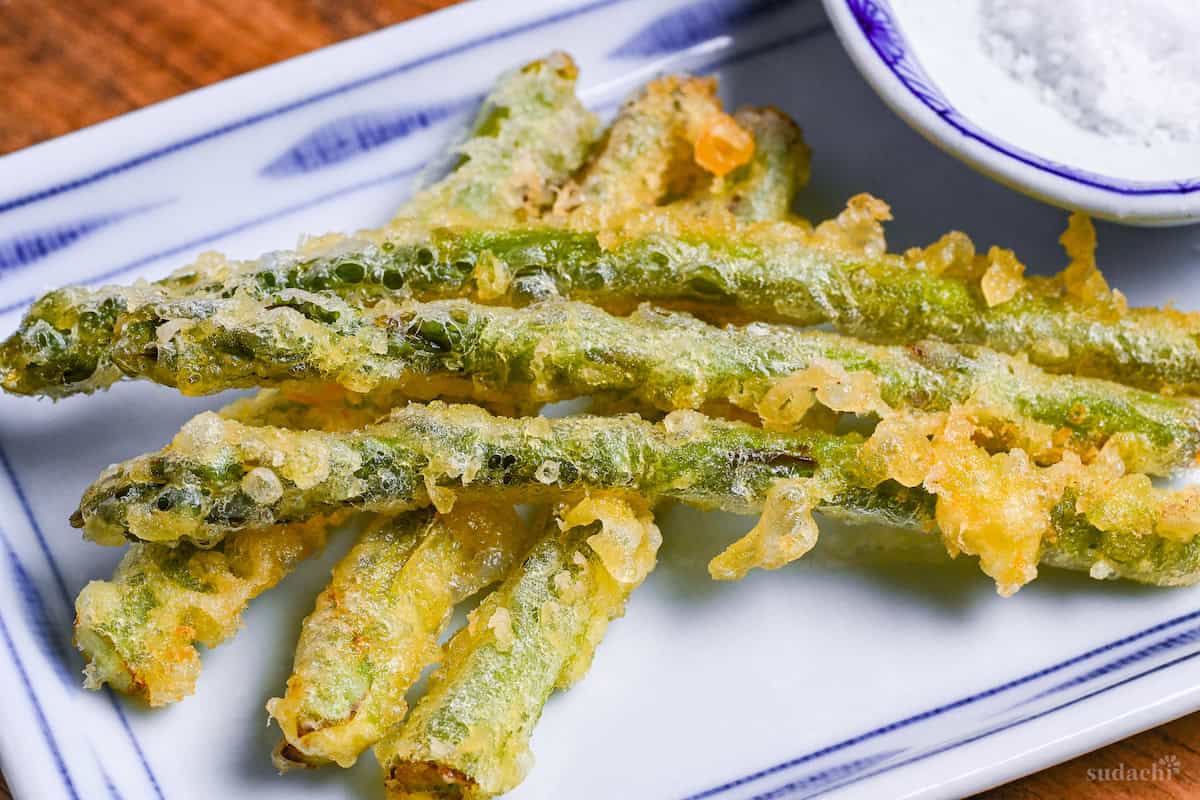 Close up of Six pieces of asparagus tempura stacked on a white plate with blue border, with a small round dish of salt next to it
