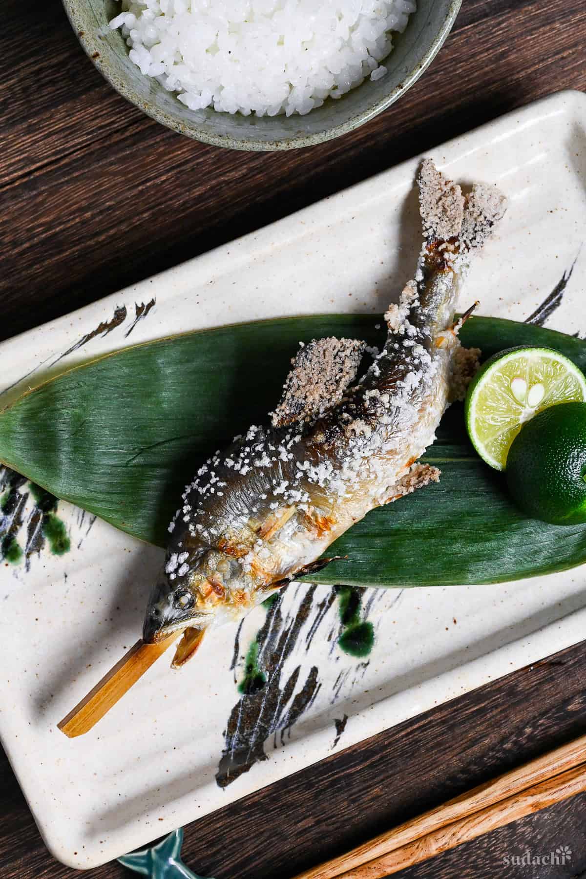 Ayu no Shioyaki (Salt Grilled Sweet Fish) on a bamboo skewer on a leaf with a halved sudachi on a rectangular ceramic plate with black and green brushstroke design