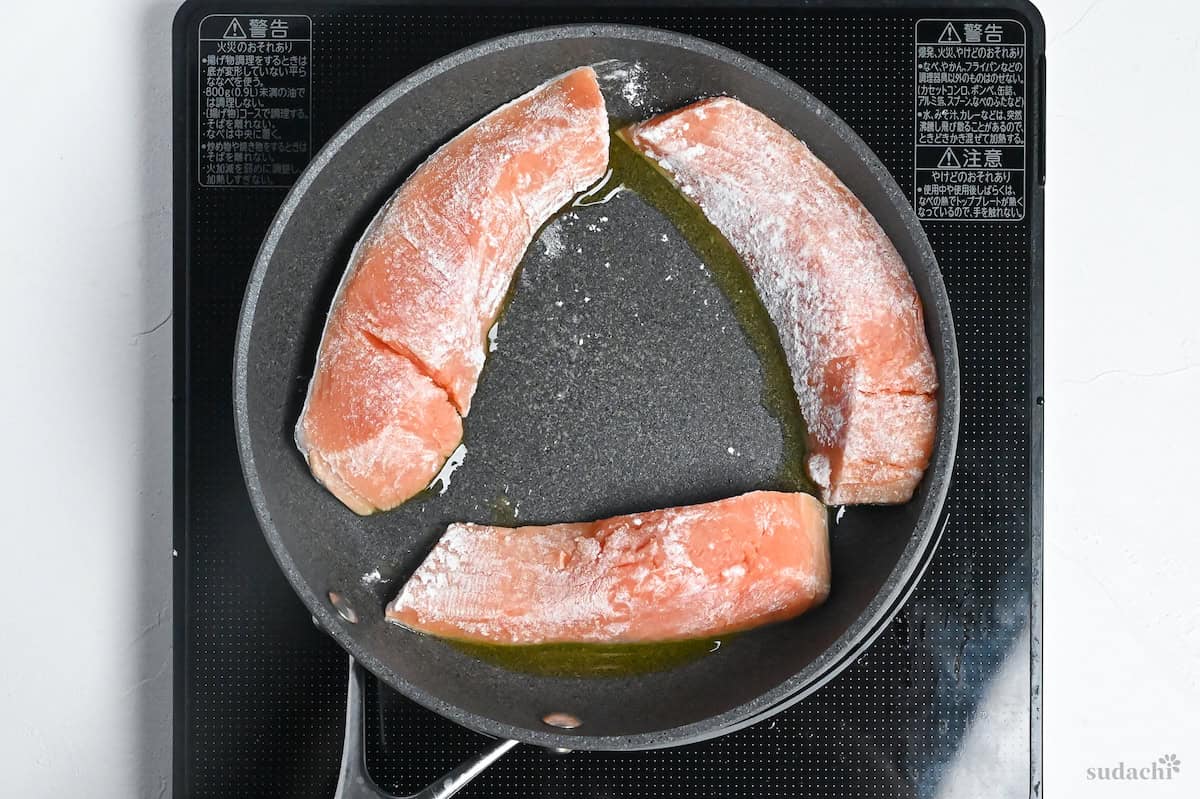 three flour-dusted salmon fillets in a frying pan with oil