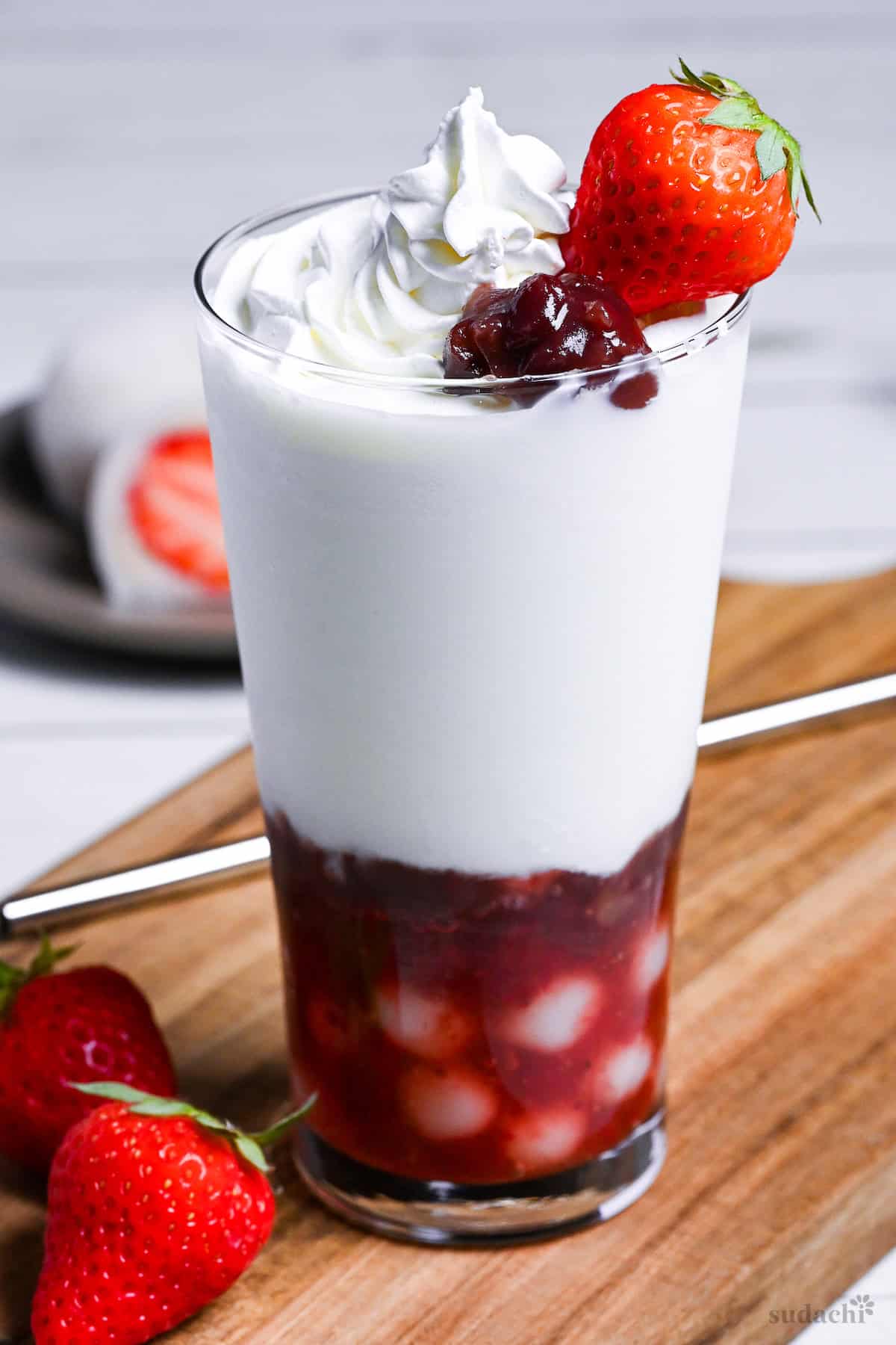 Strawberry mochi milk in a tall glass topped with whipped cream, a strawberry and boiled adzuki beans on a wooden chopping board next to ichigo daifuku on a gray plate