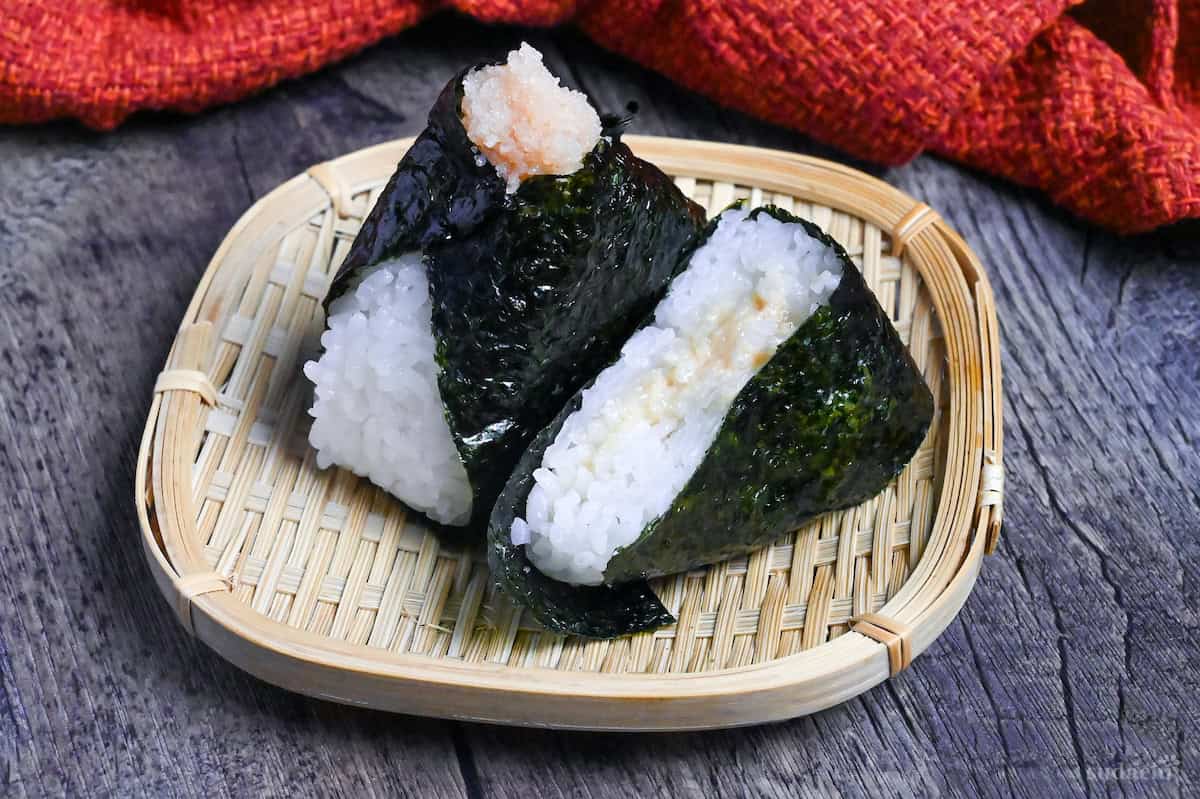 Mentaiko onigiri with one cut in half to show the filling on a small bamboo tray on a dark gray wood-effect background with red fabric in the background