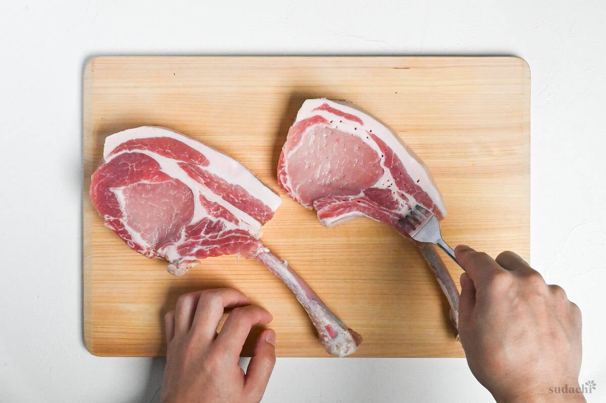 two bone-in pork chops being stabbed with a fork on a bamboo cutting board on a white background