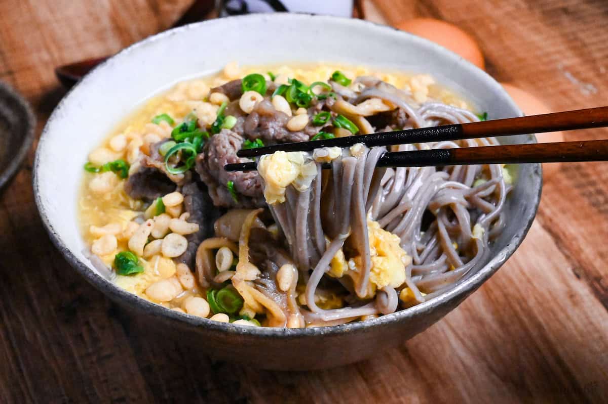Nikutama Ankake Soba (Beef & Egg Soba Noodle Soup) being picked up with chopsticks from a white and brown bowl