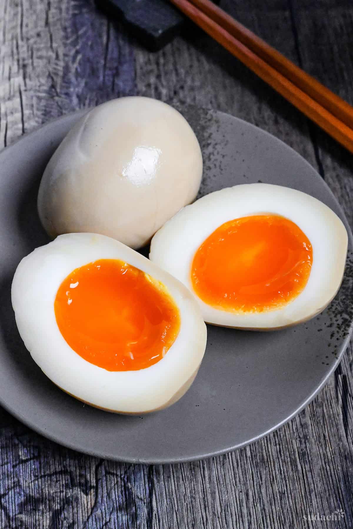 Two Ramen Eggs (Ajitsuke Tamago) with one cut in half to reveal jelly yolk on a gray plate next to wooden chopsticks on a ash-wood background
