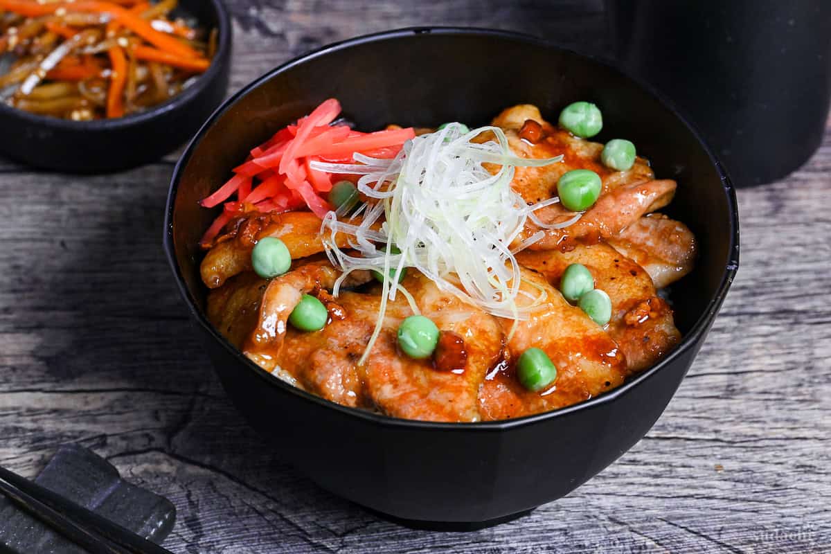 Japanese pork bowl (butadon) made with caramelized pork belly slices, green peas, pink pickled ginger and strings of Japanese leek in a black bowl on a wood-effect background