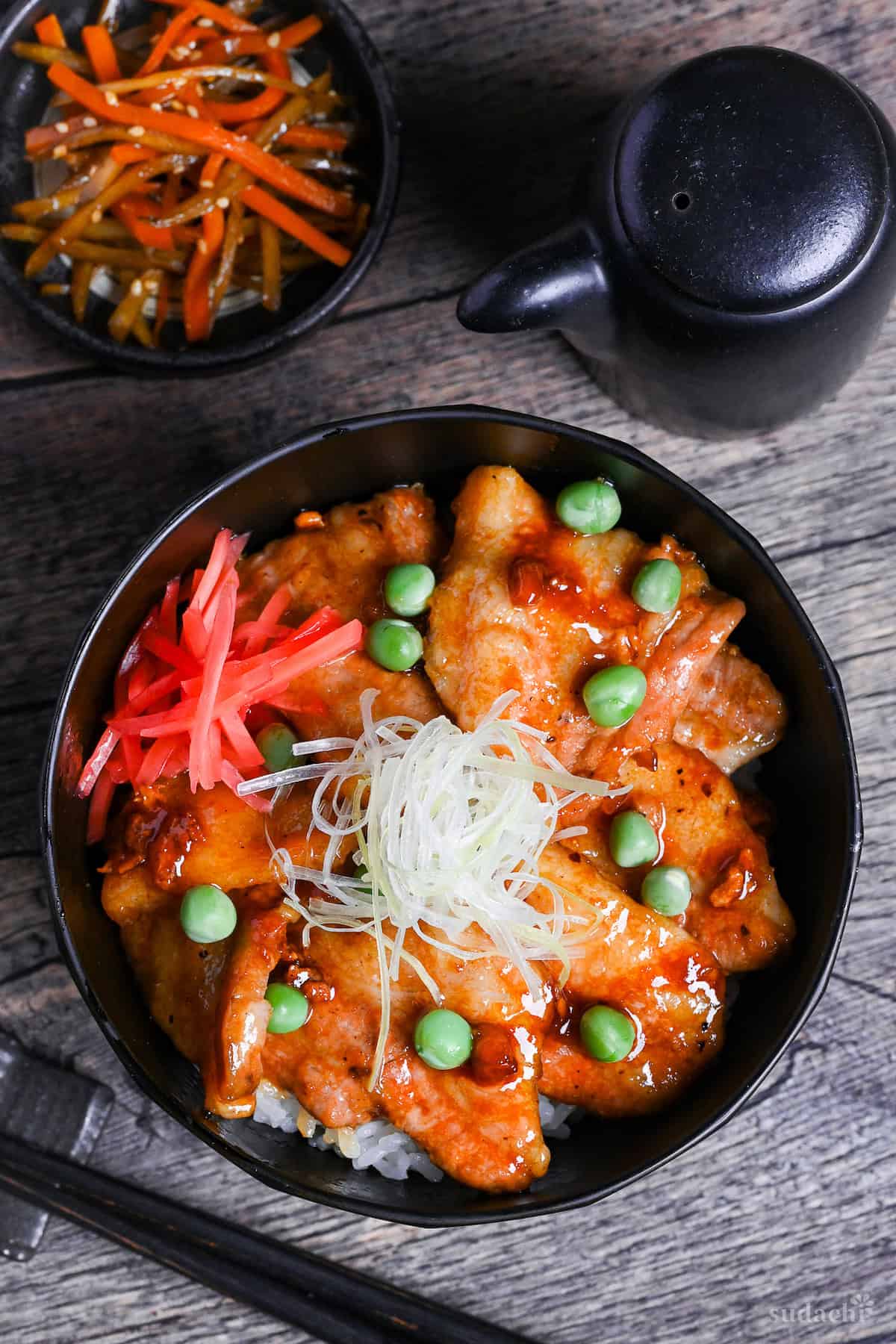 Japanese pork bowl (butadon) made with caramelized pork belly slices, green peas, pink pickled ginger and strings of Japanese leek in a black bowl on a wood-effect background top down