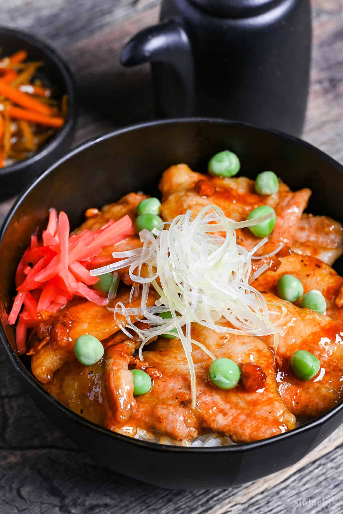 Japanese pork bowl (butadon) made with caramelized pork belly slices, green peas, pink pickled ginger and strings of Japanese leek in a black bowl on a wood-effect background