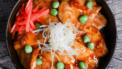 Japanese pork bowl (butadon) made with caramelized pork belly slices, green peas, pink pickled ginger and strings of Japanese leek in a black bowl on a wood-effect background