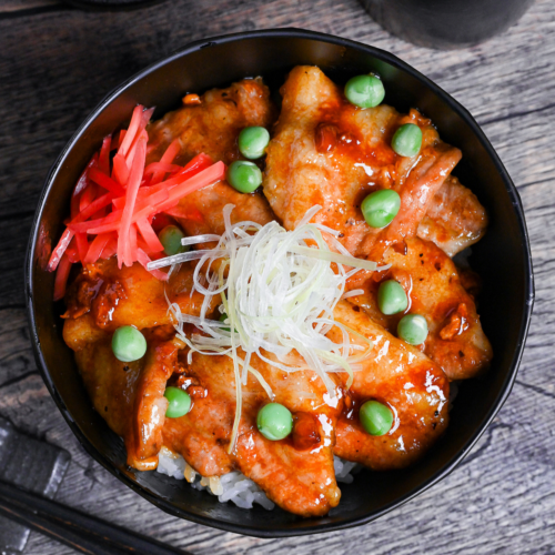 Japanese pork bowl (butadon) made with caramelized pork belly slices, green peas, pink pickled ginger and strings of Japanese leek in a black bowl on a wood-effect background